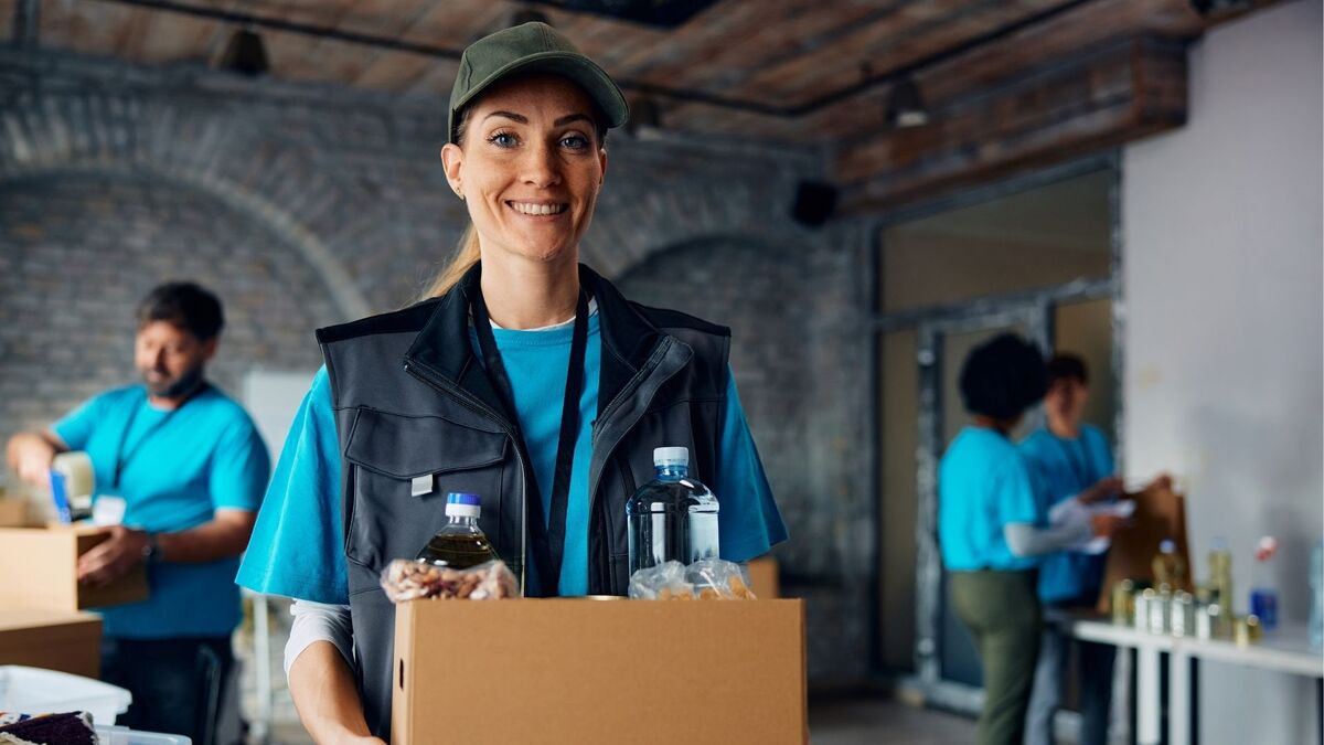 Uśmiechnięta wolontariuszka stoi na pierwszym planie i trzyma karton z produktami spożywczymi, w tym butelkami wody i innych napojów. Ma na sobie niebieską koszulkę, czarną kamizelkę, identyfikator na smyczy oraz czapkę z daszkiem. W tle widać innych wolontariuszy pakujących paczki w przestrzeni z ceglaną ścianą i drewnianym sufitem.