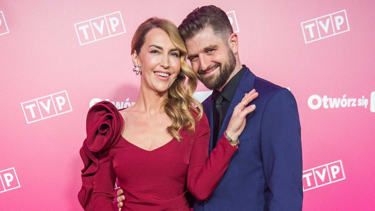 Mateusz Lisiecki-Waligórski pozuje do zdjęcia z żoną na tle ścianki z logo TVP. Kobieta ma na sobie bordową suknię, a on granatowy garnitur z czarną koszulą i krawatem. Oboje stoją blisko siebie podczas wydarzenia promocyjnego Telewizji Polskiej.