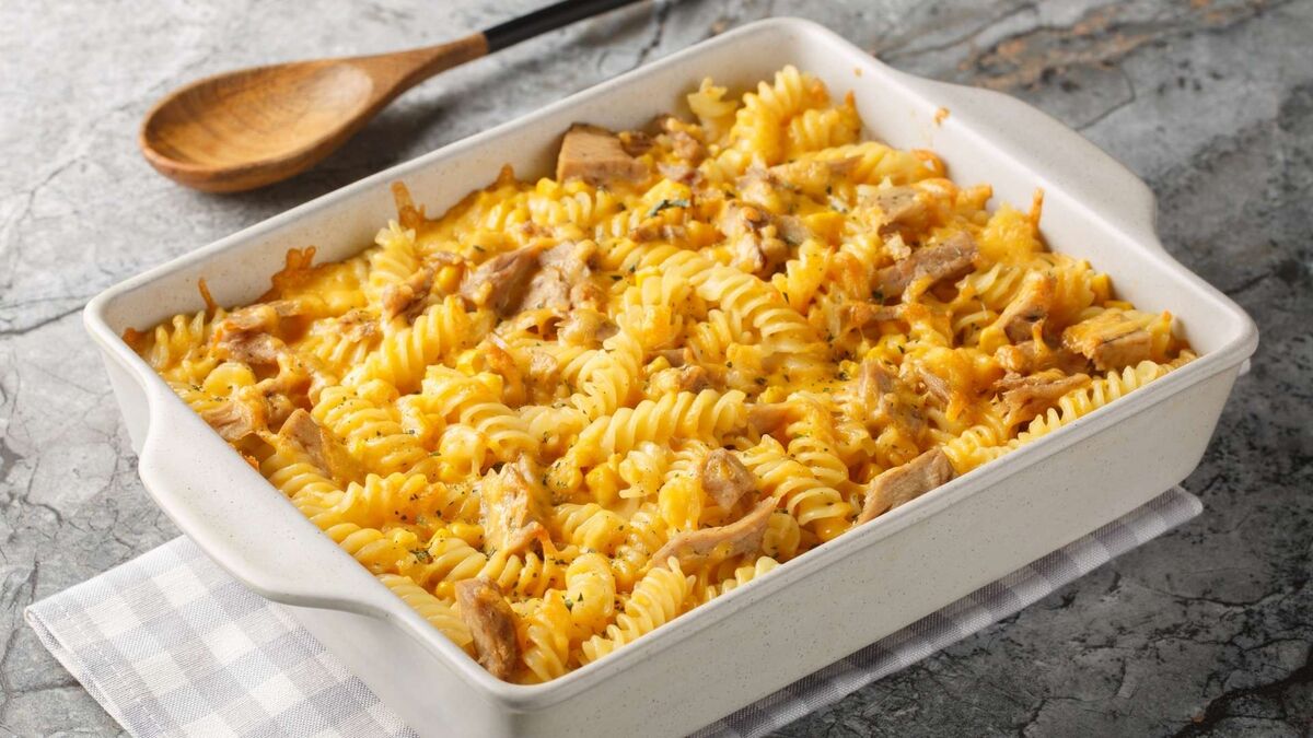 Złocista zapiekanka makaronowa z fusilli wypełnia jasne naczynie żaroodporne, a roztopiony ser tworzy apetyczną, ciągnącą się warstwę z lekko przypieczonymi brzegami. Wśród makaronu widać kawałki mięsa i delikatne zioła, które przełamują kremowy, serowy kolor dania i sugerują sycący, domowy smak. Całość stoi na szarej, kamiennej powierzchni na jasnej ściereczce, a obok leży drewniana łyżka, jakby zapiekanka dopiero co została wyjęta z piekarnika i czekała na podanie.