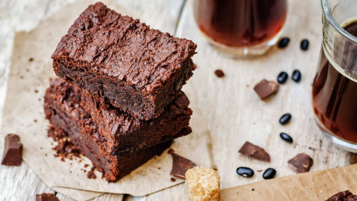 Kawałki brownie z fasoli są ułożone jeden na drugim na jasnym, drewnianym blacie. Ciasto ma ciemny, czekoladowy kolor oraz zwartą, lekko wilgotną strukturę z wyraźnie zaznaczoną, nierówną powierzchnią. Wokół leżą fragmenty czekolady, pojedyncze ziarna czarnej fasoli oraz kostki brązowego cukru. W tle widoczne są szklanki z ciemnym napojem, a całość tworzy prostą, rustykalną kompozycję.