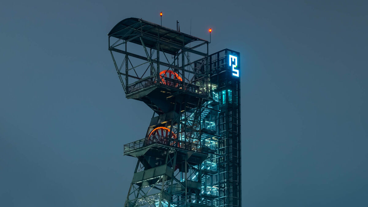 Ujęcie przedstawia konstrukcję wieży szybu górniczego o zmroku, z widocznymi kołami linowymi i platformami technicznymi. Po prawej stronie znajduje się podświetlany szyb windowy z jasnymi schodami i windą. Na górze świeci znak „MŚ”, a tło stanowi jednolite, ciemnoniebieskie niebo.
