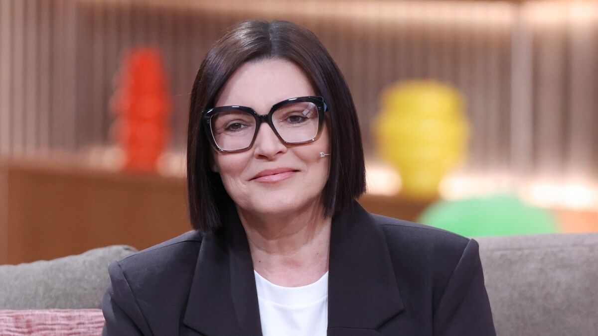Ilona Felicjańska siedzi na kanapie i patrzy przed siebie z delikatnym uśmiechem. Ma krótkie, ciemne włosy, duże czarne okulary, czarną marynarkę założoną na białą bluzkę oraz mikrofon przy policzku. W tle widoczne są rozmyte elementy studia z kolorowymi dekoracjami i pionowymi lamelami.