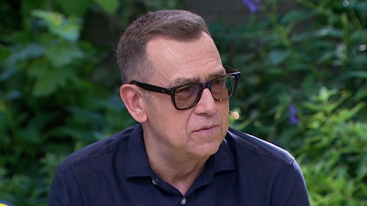 Maciej Orłoś siedzi w plenerze, ubrany w ciemnogranatową koszulę i okulary z przyciemnianymi szkłami. Ma przy twarzy niewielki mikrofon i spogląda w bok z poważnym wyrazem twarzy. W tle widać gęstą zieloną roślinność z pojedynczymi fioletowymi kwiatami.