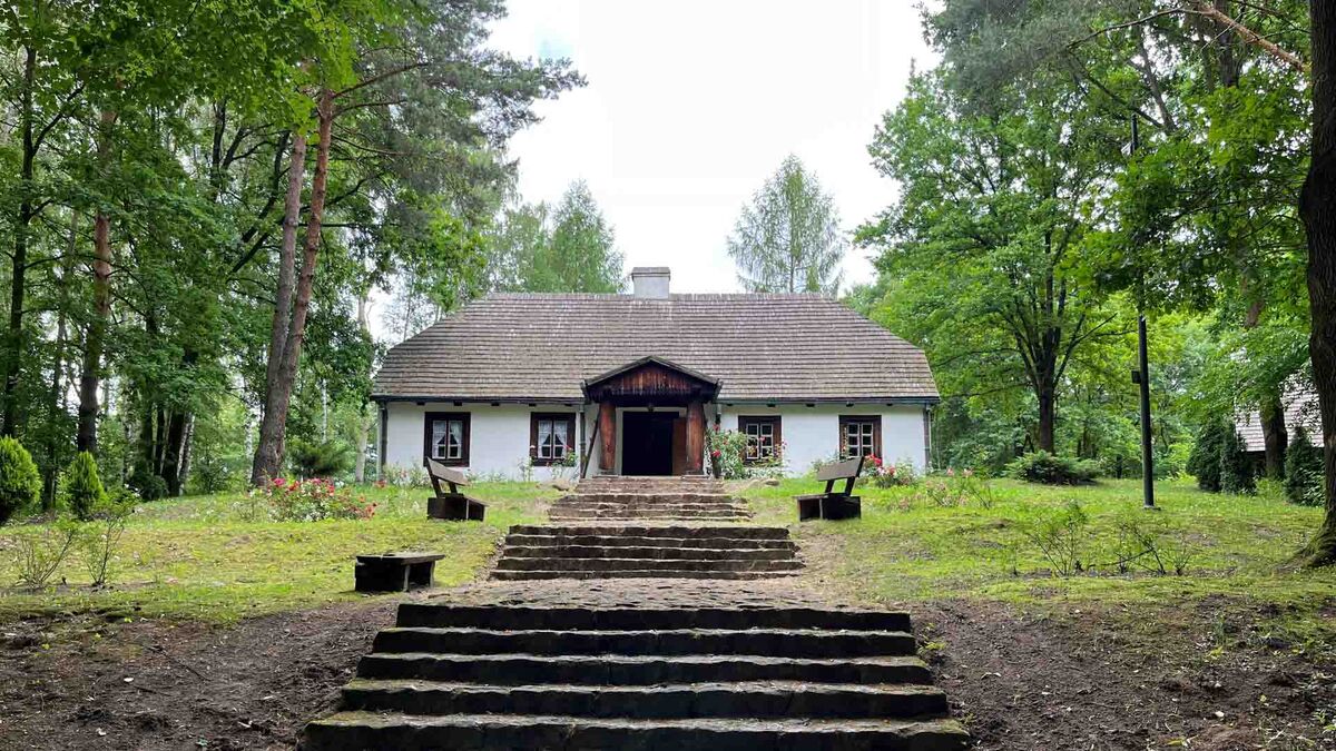Na zdjęciu widoczny jest zabytkowy drewniany dwór otoczony drzewami i zielenią. Do wejścia prowadzą kamienne schody, a po obu stronach ścieżki ustawiono ławki i kwitnące krzewy. Budynek znajduje się na terenie parku z wysokimi drzewami.