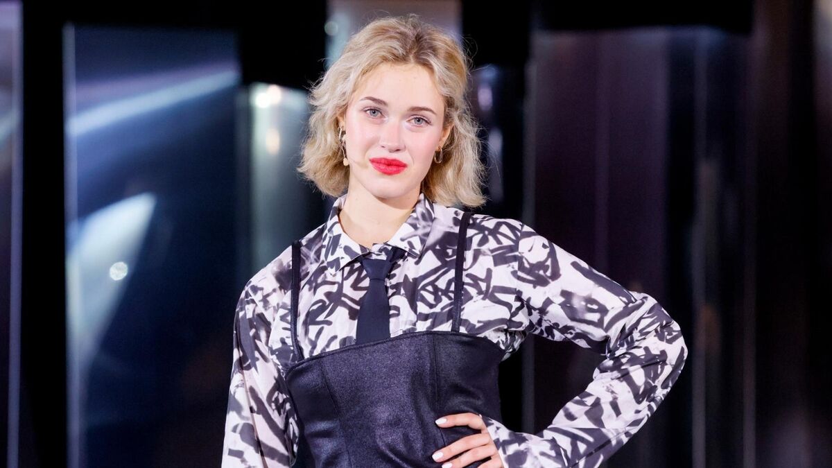 Helena Englert stoi na scenie i pozuje z ręką opartą na biodrze. Ma krótkie, falowane blond włosy oraz wyrazisty makijaż z czerwonymi ustami. Ubrana jest w koszulę w czarno-biały wzór z krawatem oraz czarny gorset na ramiączkach, a w tle widać ciemną scenografię i oświetlenie sceniczne.