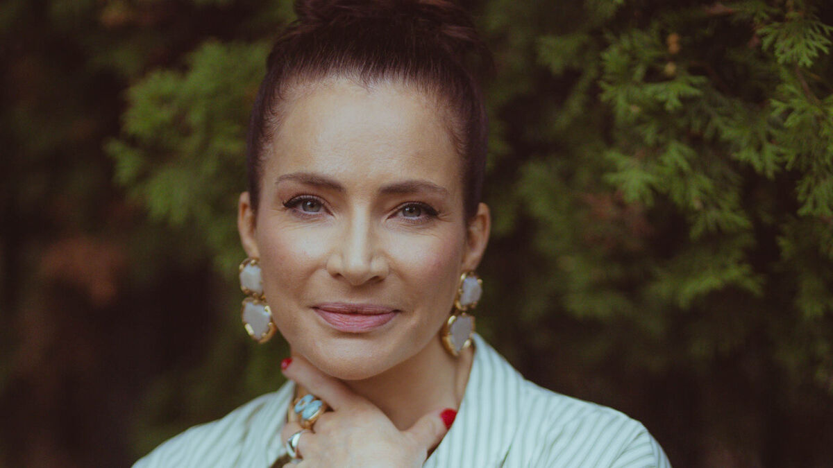 Anna Dereszowska z serialu „Felicita” ma włosy upięte wysoko i duże ozdobne kolczyki. Aktorka opiera dłoń pod brodą, a na palcach ma kilka pierścionków. W tle widoczne są gęste zielone krzewy.
