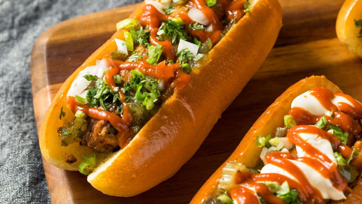 Wege hot dog podany jest w miękkiej bułce i obficie udekorowany dodatkami. Na wierzchu widoczne są kawałki cebuli, ogórków konserwowych, świeże zioła oraz sosy w jasnym i czerwonym kolorze. Danie leży na drewnianej desce, a obok znajduje się drugi hot dog przygotowany w podobny sposób.